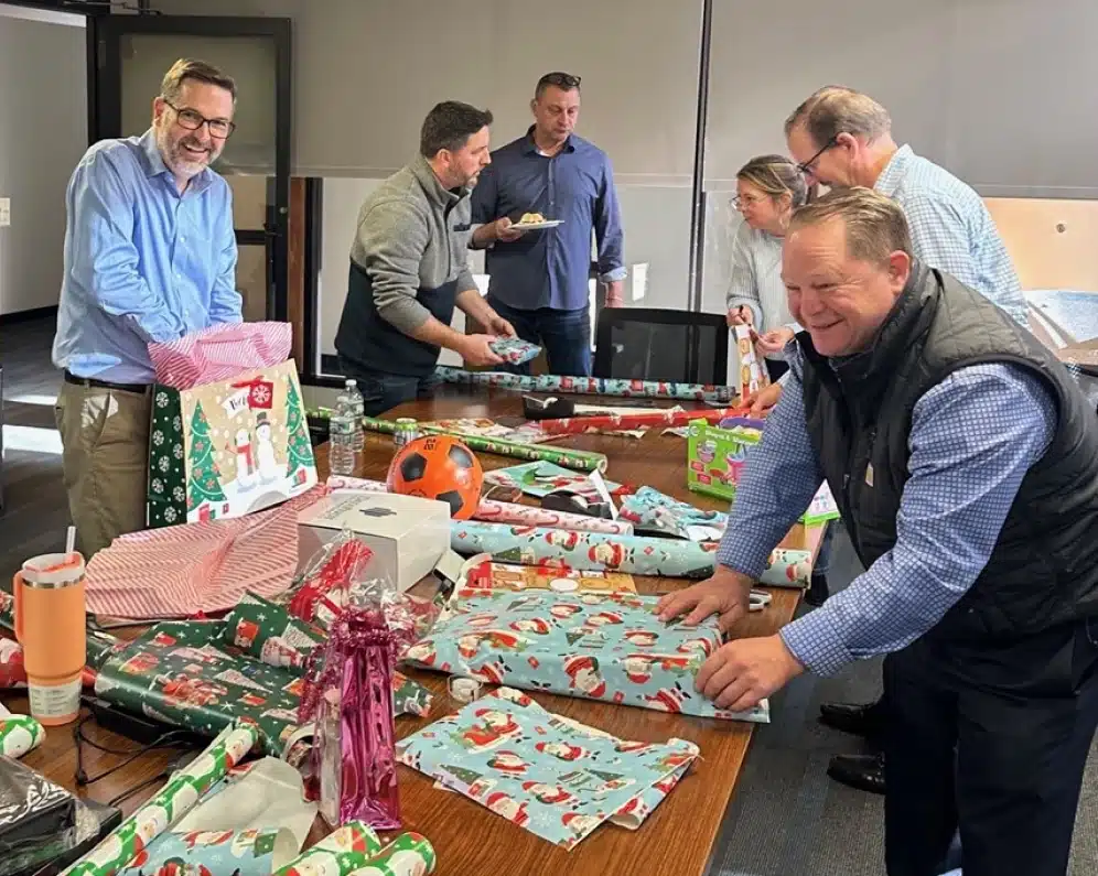 Employees wrapping holiday gifts for a charitable volunteer project inside a modern office meeting room