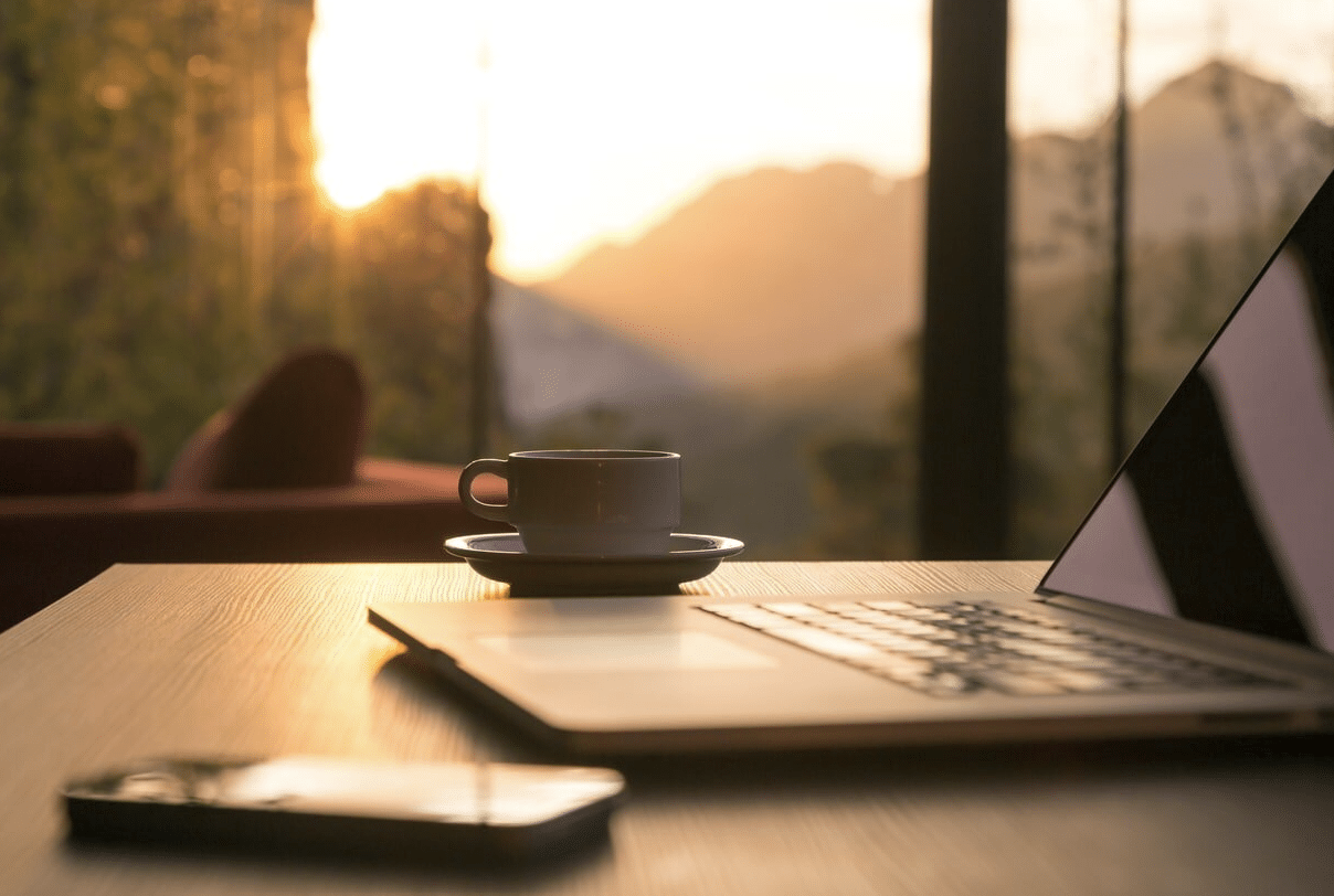 Cup of coffee on wooden desk beside laptop and phone, with sunrise through window background