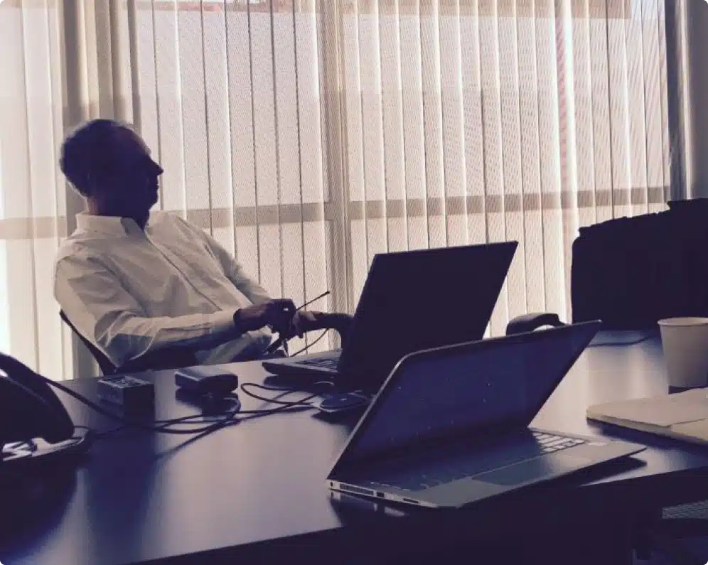 Business professional seated at table with blinds drawn, surrounded by laptops and work tools.